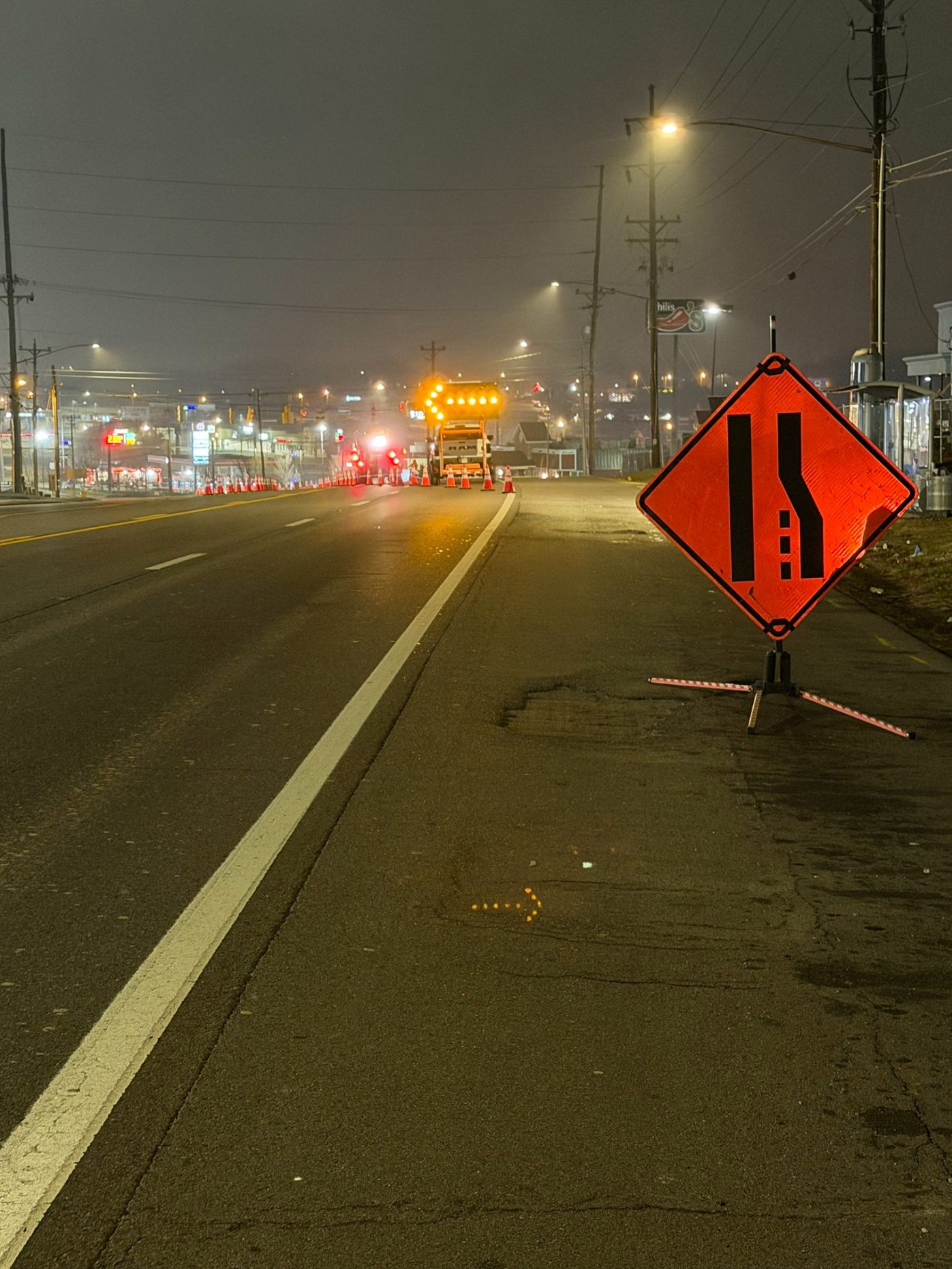 Lane-reduction warning sign in the foreground with an arrow board and cone taper beyond it at night