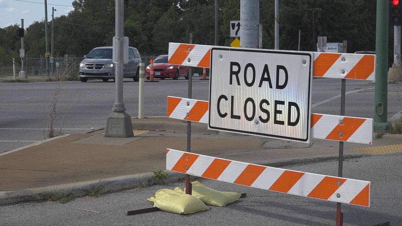 Road Closed sign mounted on an orange and white Type III barricade, weighted with sandbags at an intersection