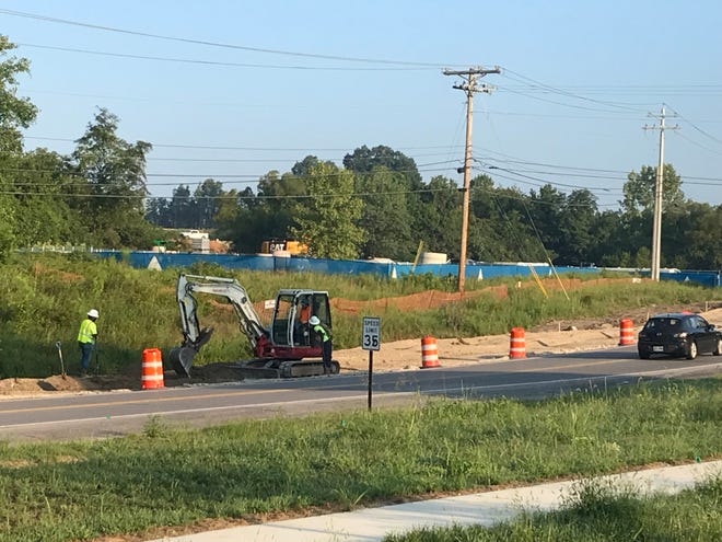Mini excavator and crew in high-visibility vests working along the shoulder of a 35 mph road, separated from traffic by orange drums